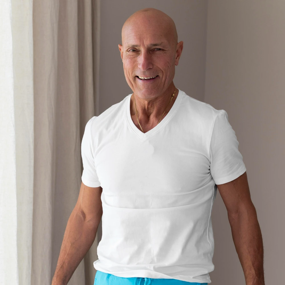 Smiling bald man wearing a white v-neck t-shirt and blue shorts standing indoors.