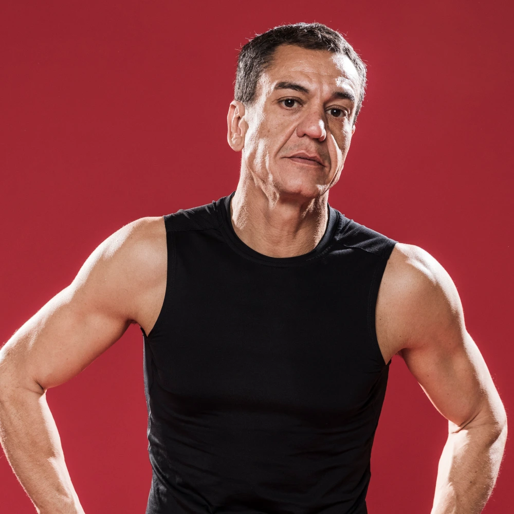 Fit middle-aged man in a black sleeveless shirt standing with hands on hips against a red background.