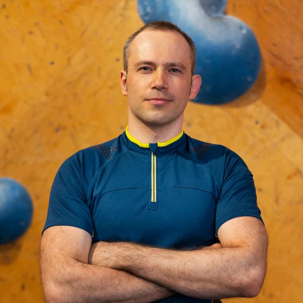 Man with short hair wearing a blue athletic shirt, standing with arms crossed in front of an orange climbing wall with large blue holds.