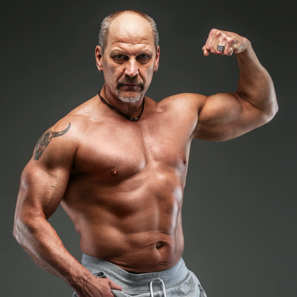 Muscular middle-aged man flexing his right arm, showing a tattoo on his left shoulder against a dark background.