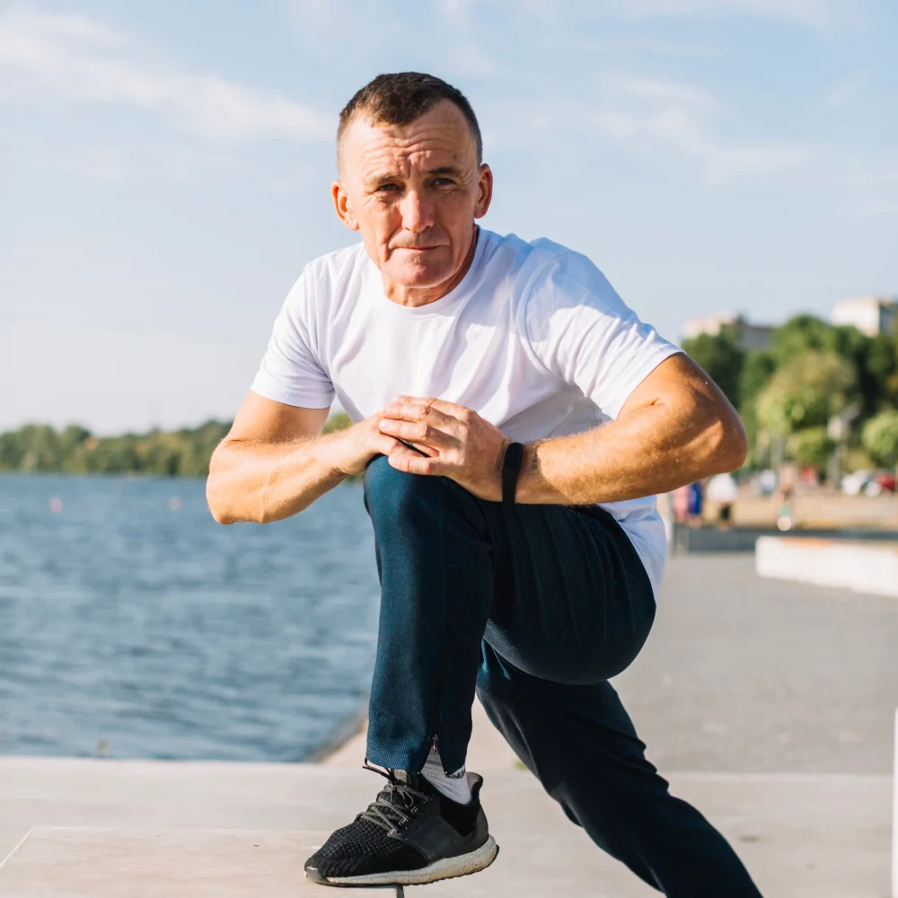 Man in white t-shirt and black sneakers stretching one leg outdoors near a body of water.