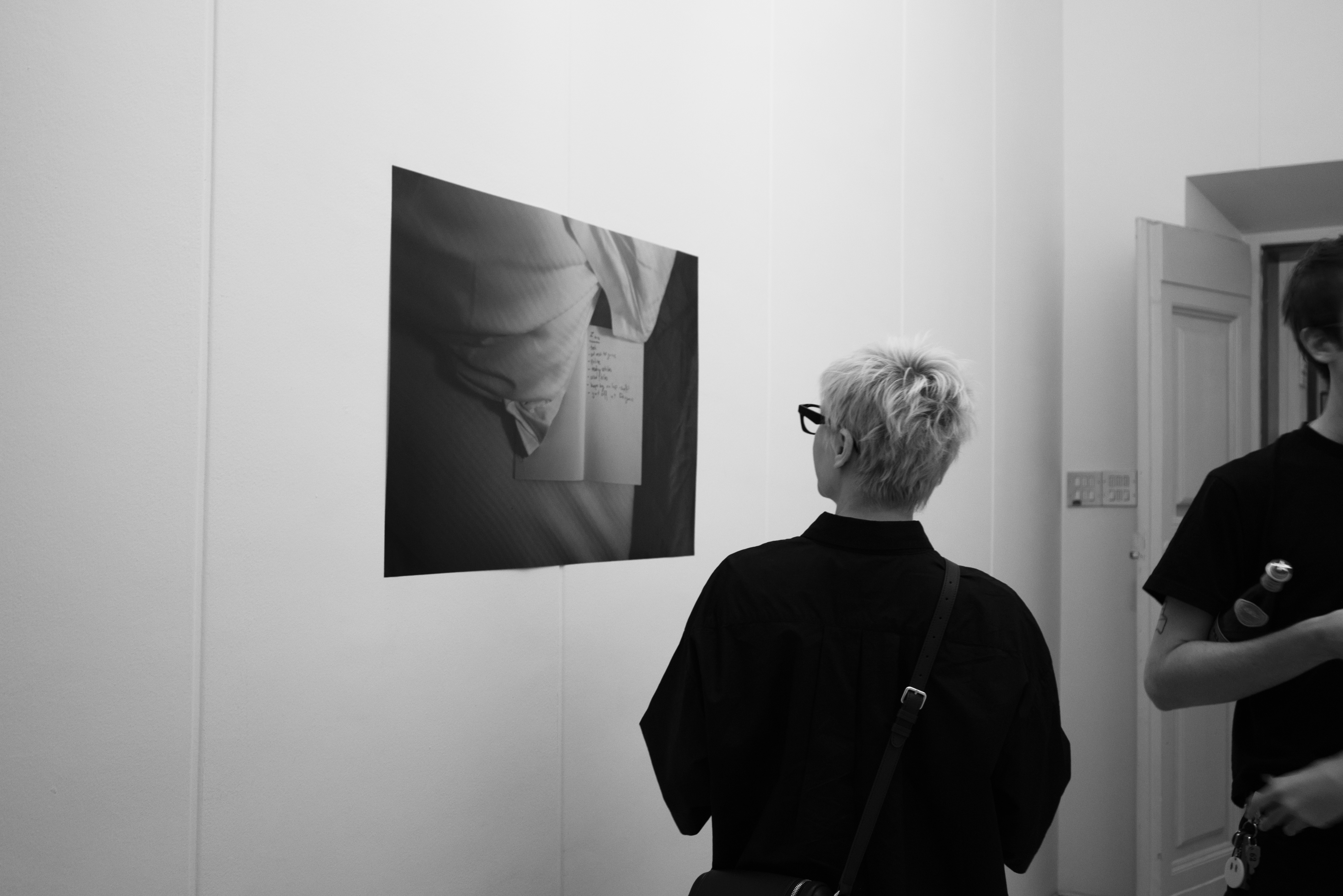 Black and White image: Hallway with photographs and person looking at photograph