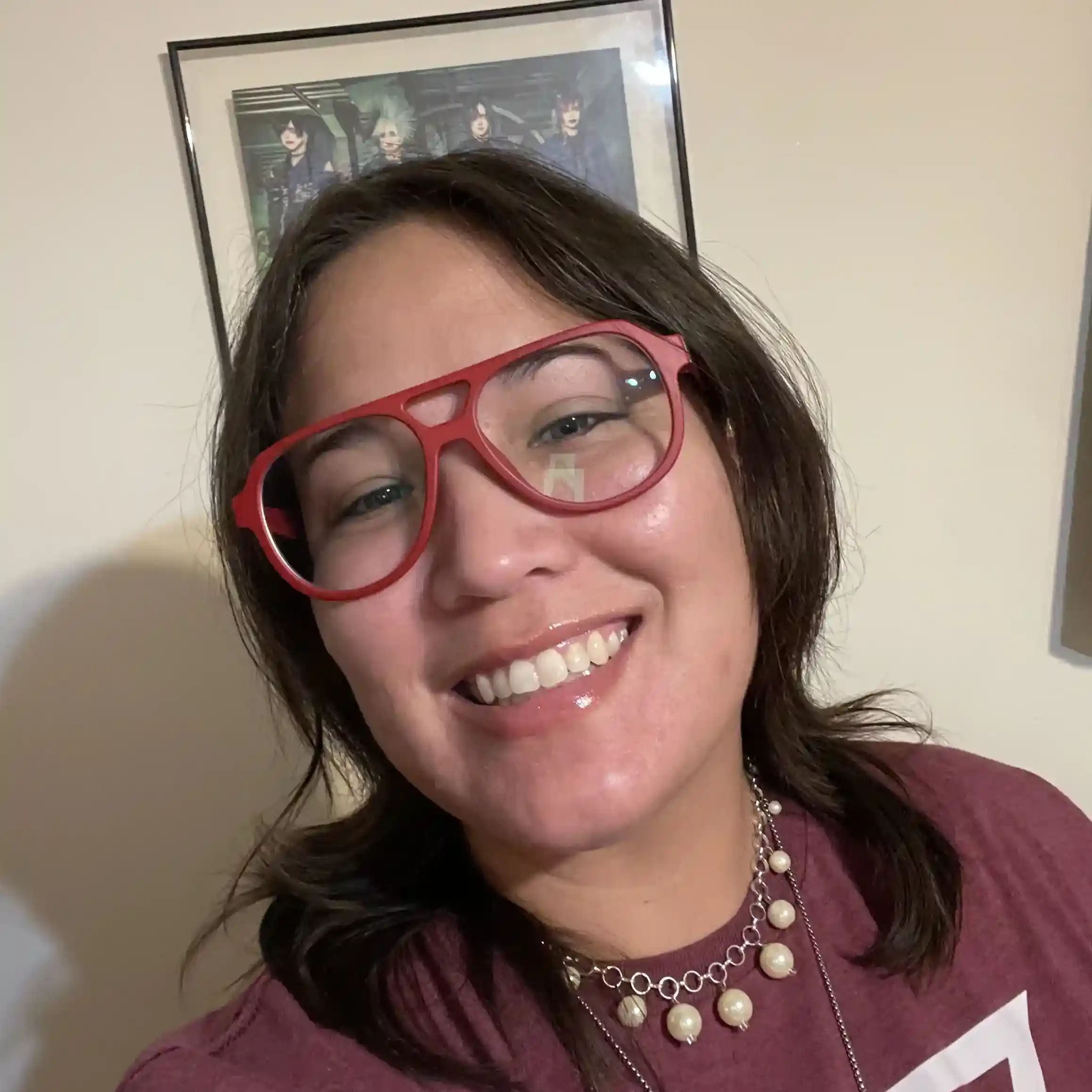 Smiling woman wearing large red glasses and a maroon shirt, standing in front of framed art.