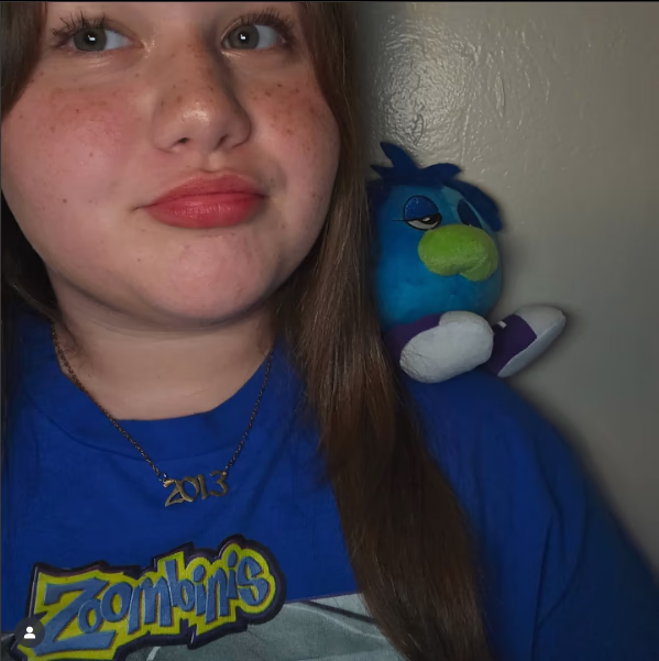 Girl wearing a blue Zoombinis shirt with a Zoombini plush on her shoulder, looking off to the side.