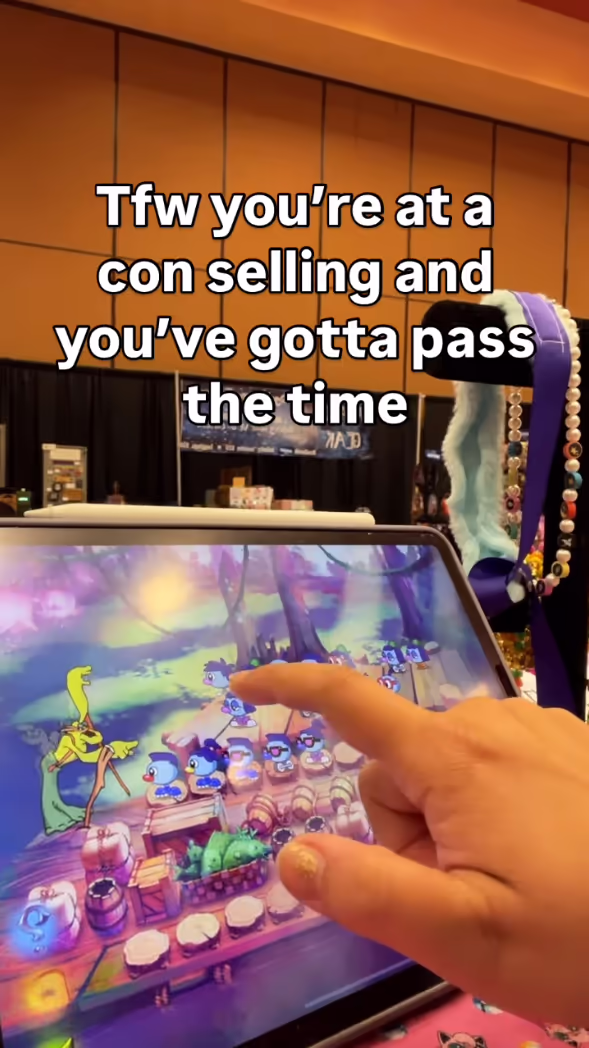 Hand pointing at Zoombinis gameplay on a tablet at a convention booth with crafts in the background.