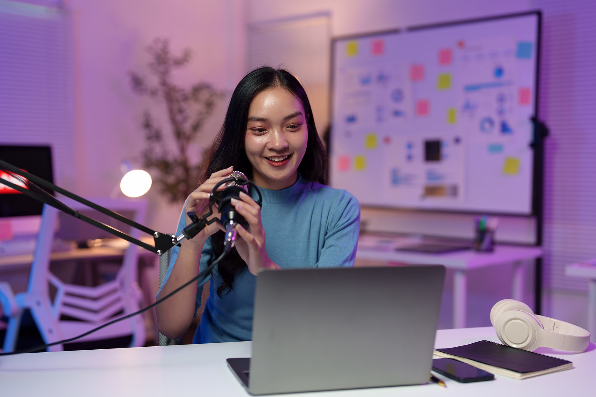 Smiling woman speaking into a microphone while recording at her desk with a laptop and headphones.