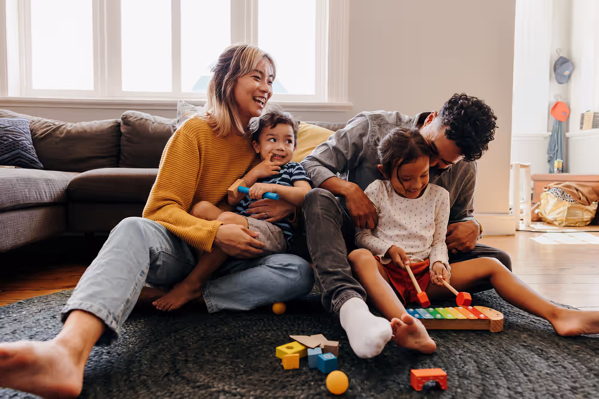 Happy family sitting on the floor playing with toys, including a rainbow xylophone and building blocks.