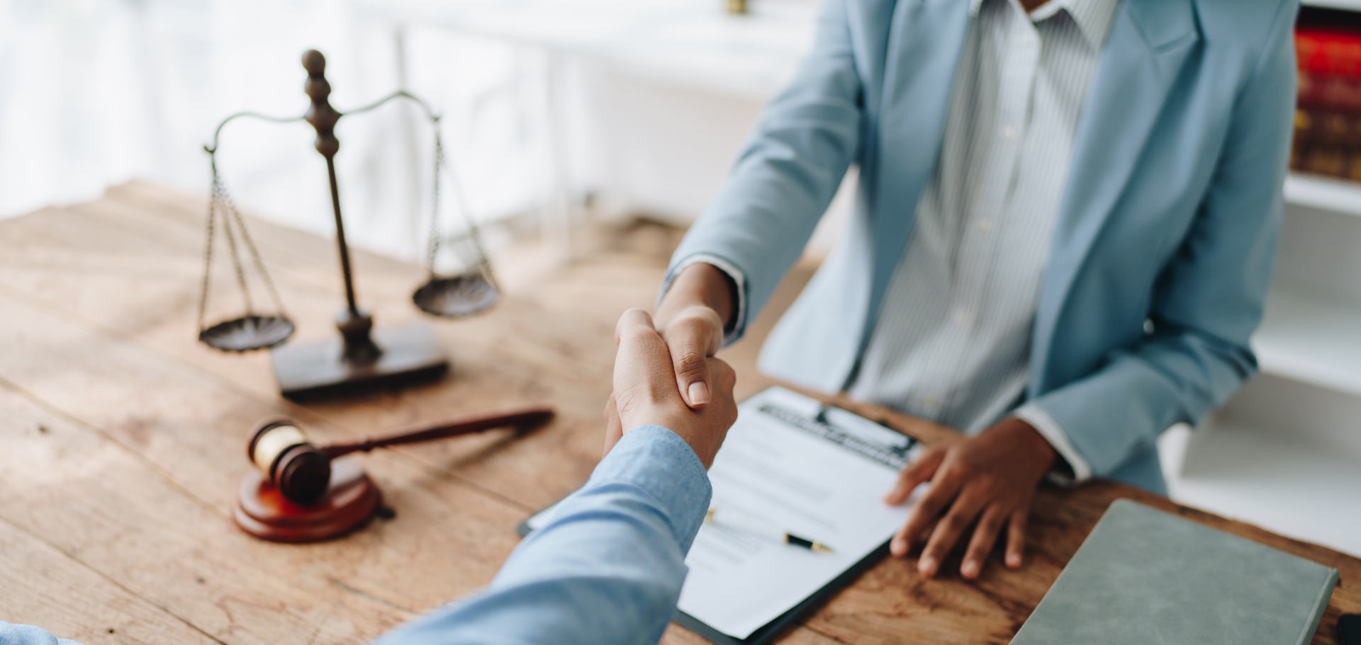 Four disabled people of color gather around a table during a meeting. A Black woman sitting on a couch gestures and speaks while the three others (a South Asian person sitting in a wheelchair, a Black non-binary person sitting in a chair, and a Black non-binary person standing with a clipboard and cane) face her and listen.