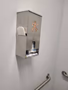 Wall-mounted stainless steel feminine hygiene dispenser in a restroom with sanitary products visible inside.