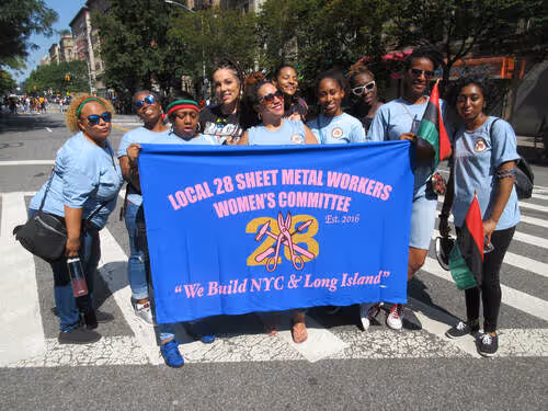 SMART Local 28 Women’s Committee members standing outdoors holding a blue banner that reads 'We Build NYC & Long Island' during a parade.