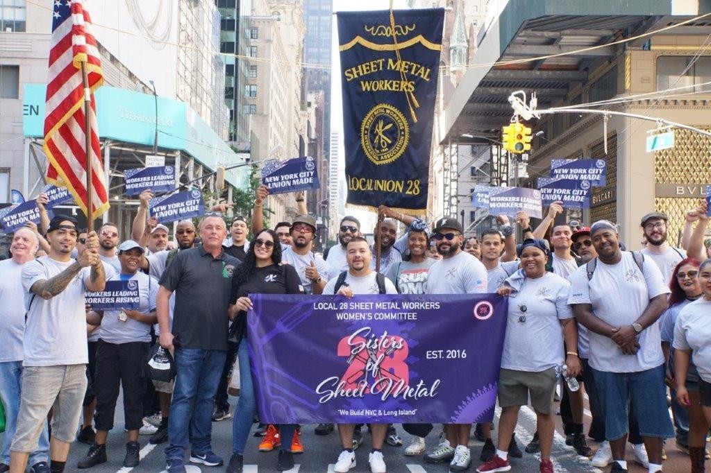 Group of SMART Local 28 sheet metal workers standing together in a city street holding banners and a “Sisters of Sheet Metal” sign at a parade or labor event.