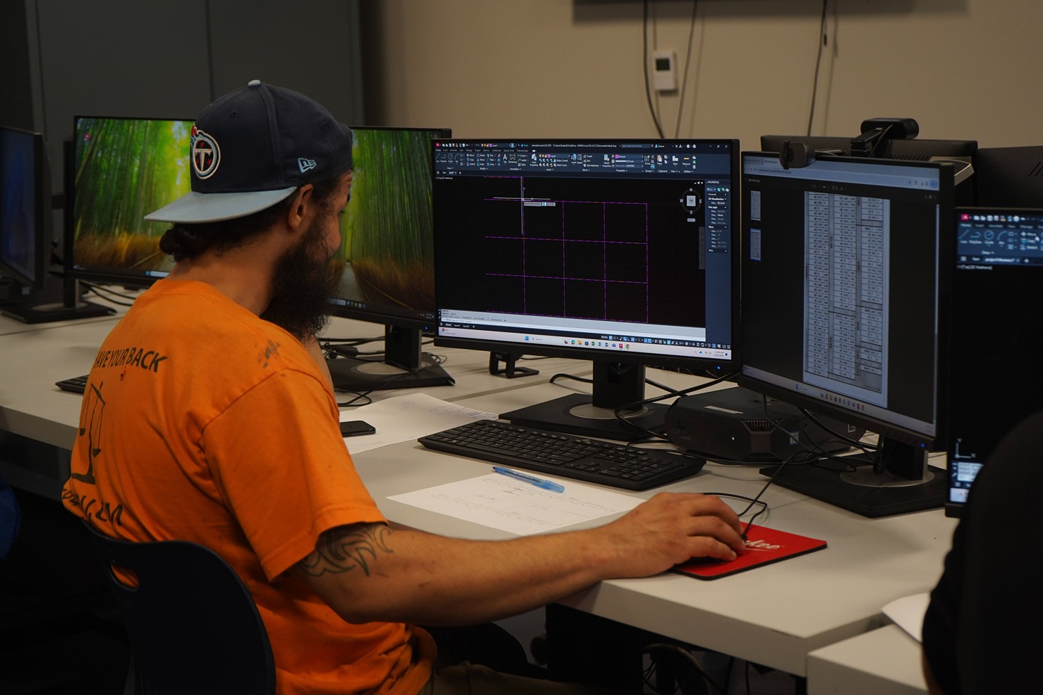 Bearded man wearing a backward baseball cap and orange shirt working on a dual-monitor desktop, viewing a grid layout on one screen and a table on the other.