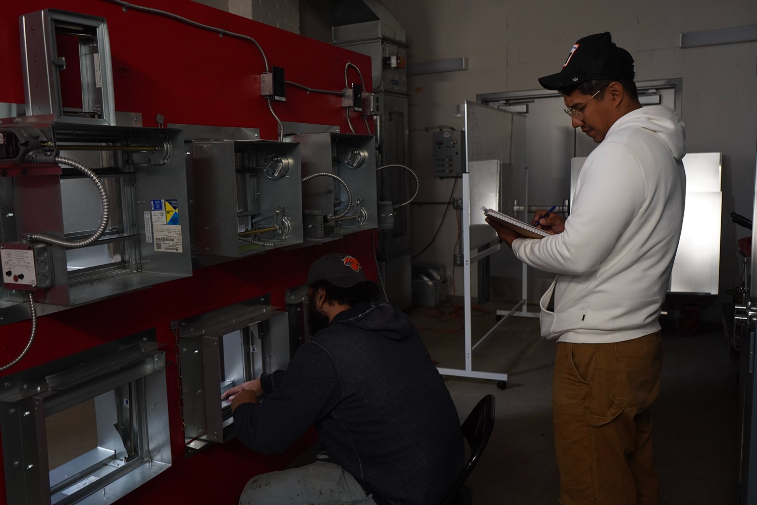 Two men in a workshop; one is kneeling and working on metal duct components mounted on a red board, the other is standing and writing notes on a clipboard.