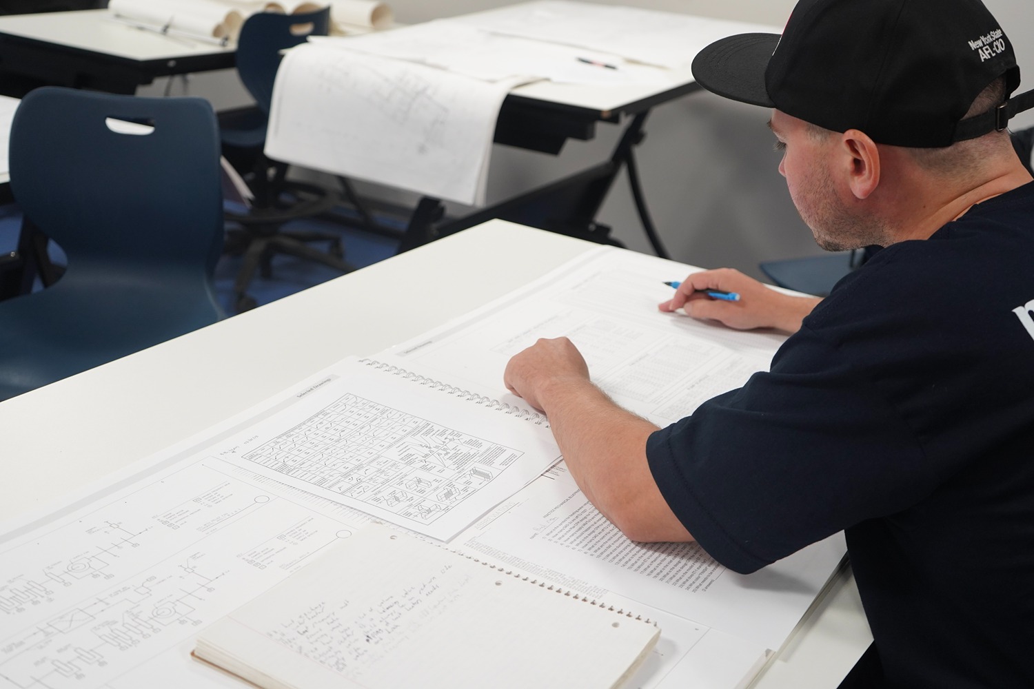 Man in a black cap and shirt studying technical drawings and notes on a white table.
