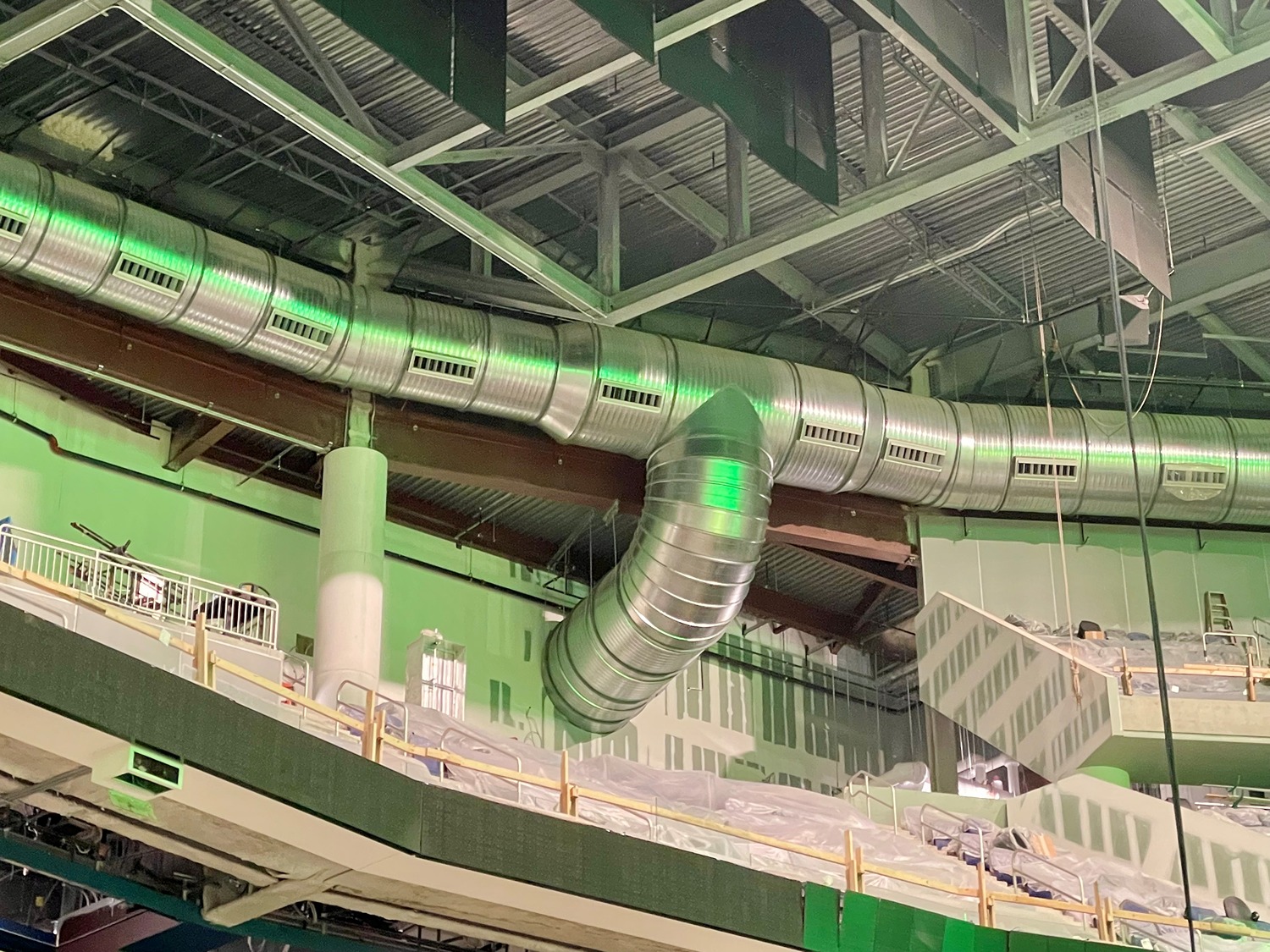 Large silver industrial ventilation pipe with green lighting mounted along an arena ceiling above covered seating areas.