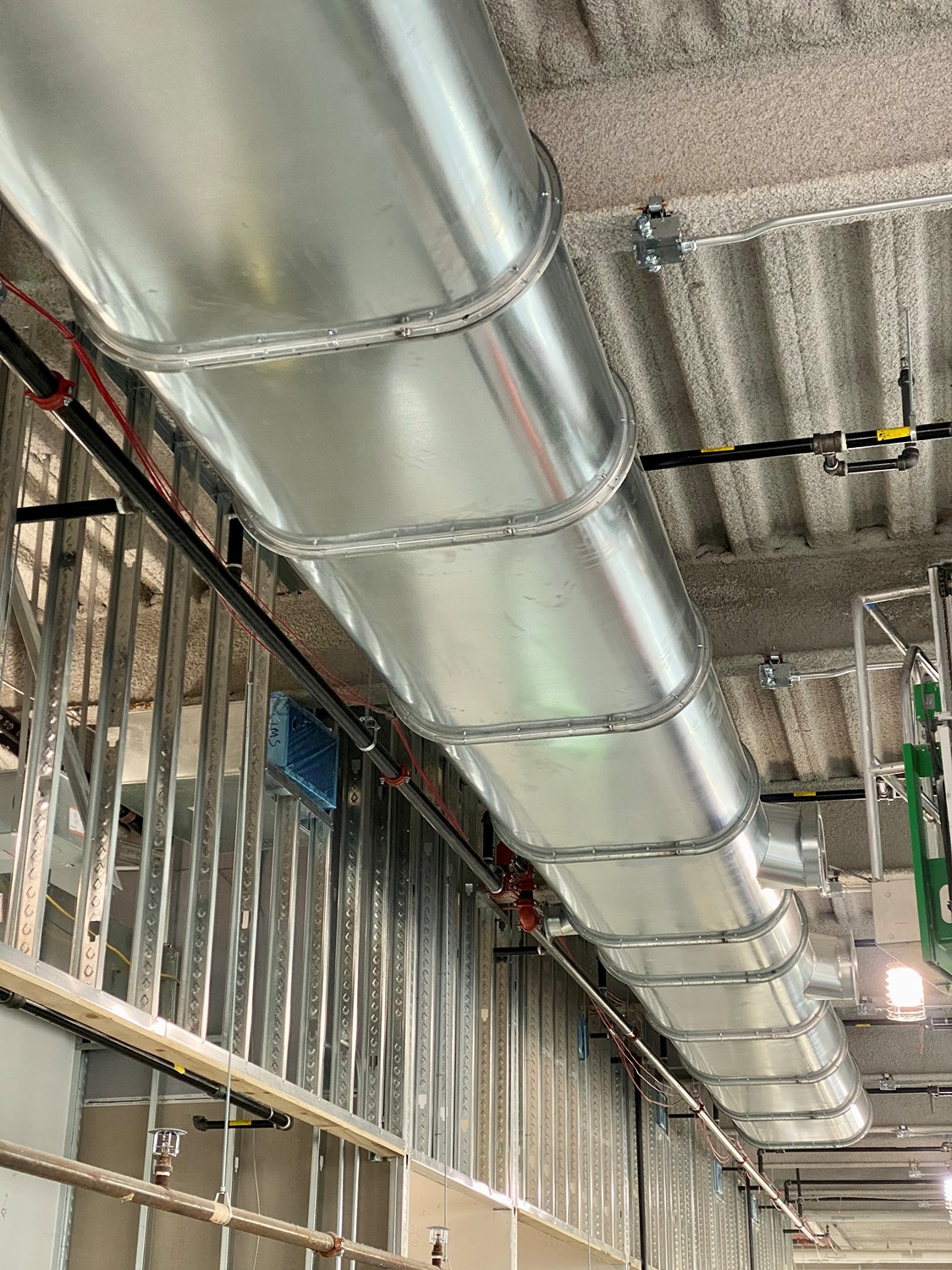 Large oval-shaped metal ductwork installed on a ceiling with exposed metal wall framing and electrical conduit.