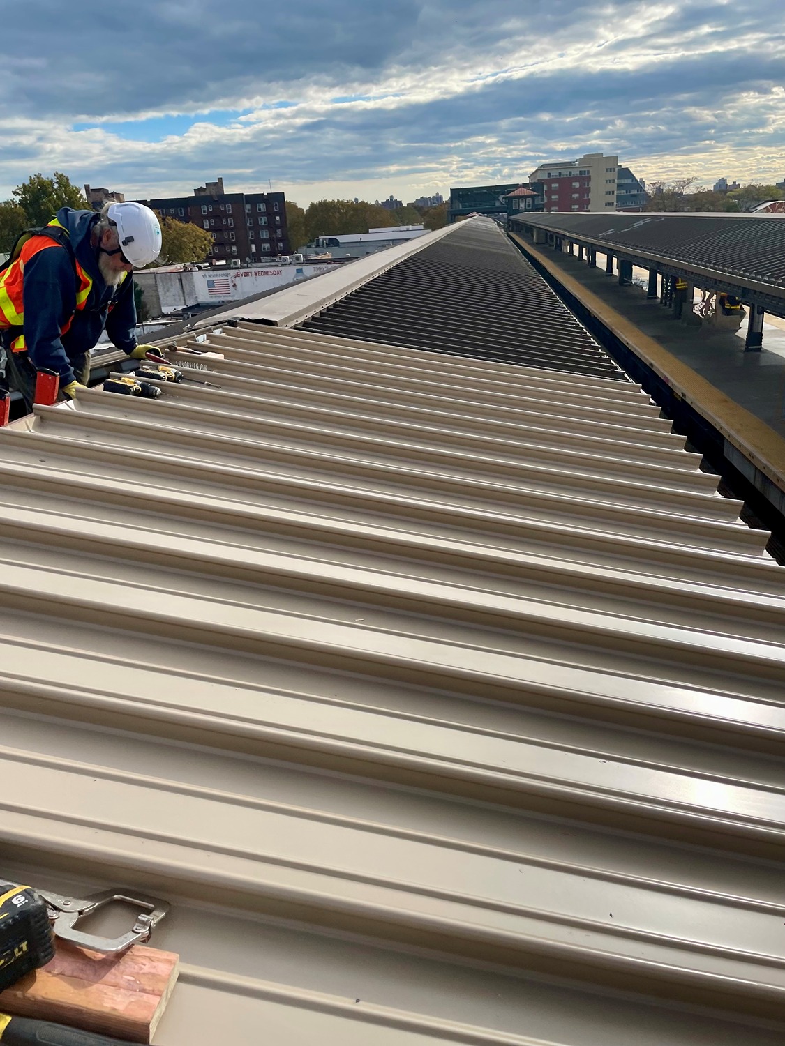 Construction worker in a safety vest and helmet working on installing metal roofing panels on a rooftop next to a train station platform.