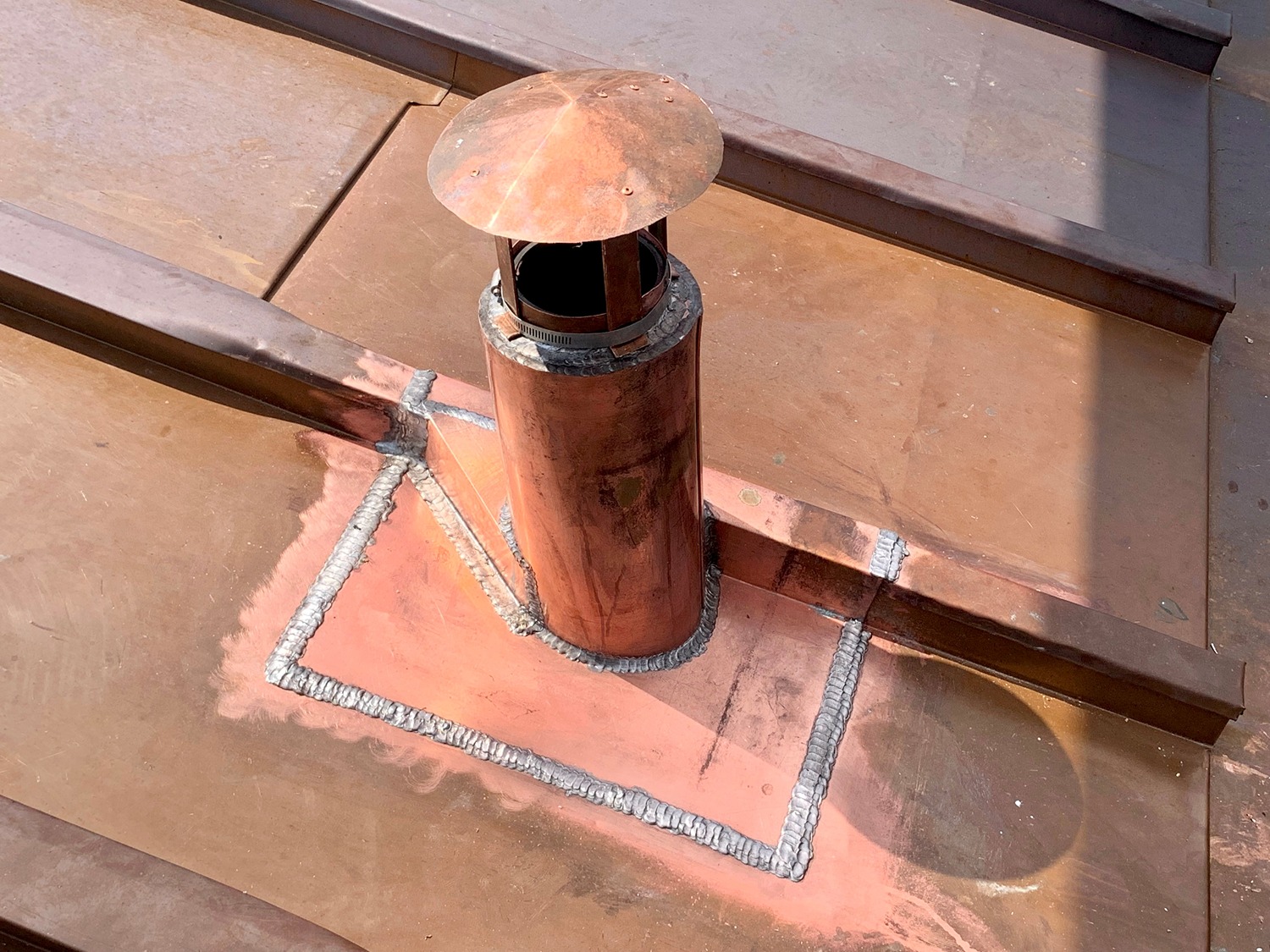 Copper chimney pipe with a round cap installed on a copper roof with visible welded seams.
