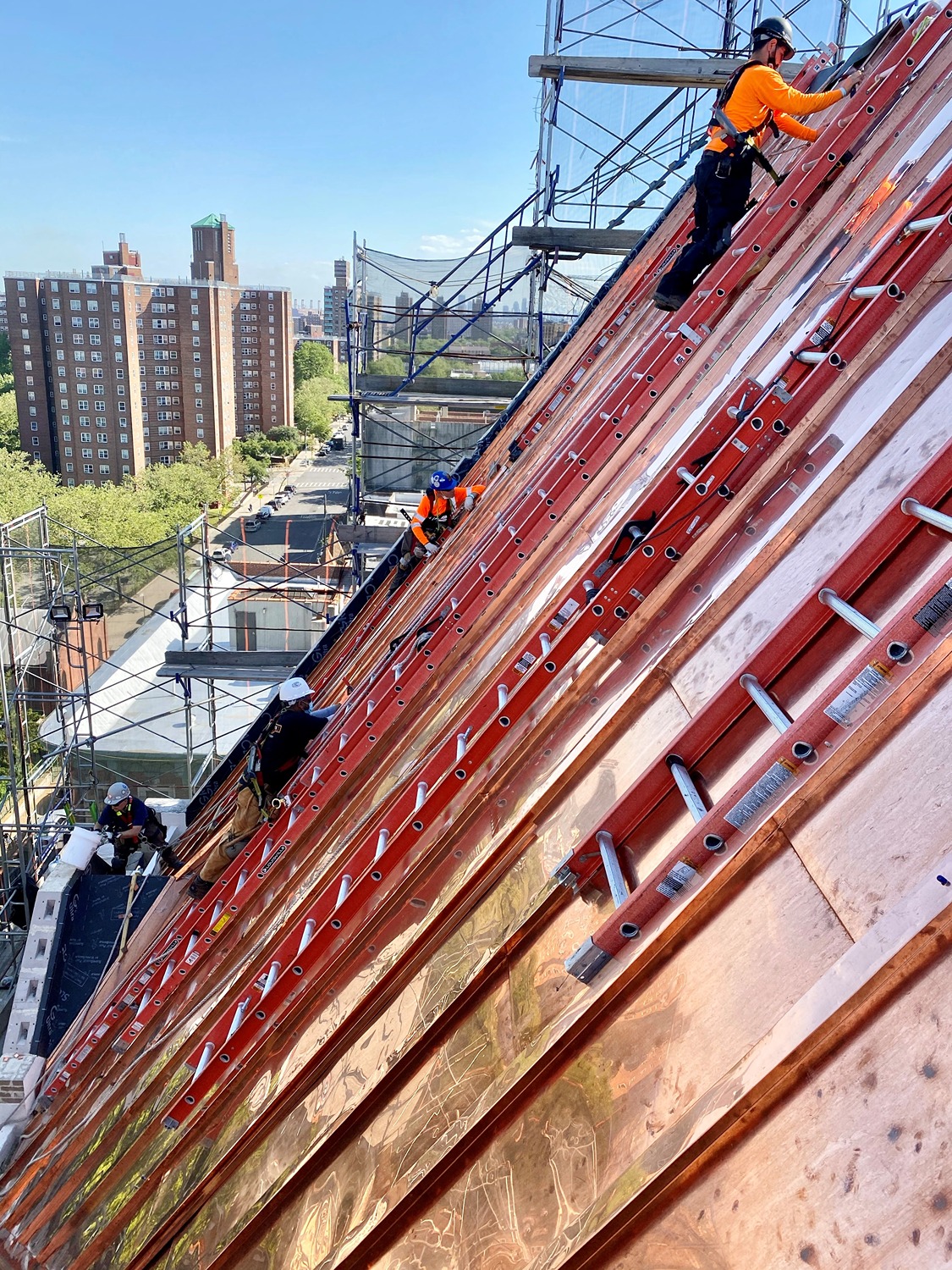 Construction workers in safety gear working on scaffolding and ladders on a steep roof in an urban area with buildings and trees in the background.