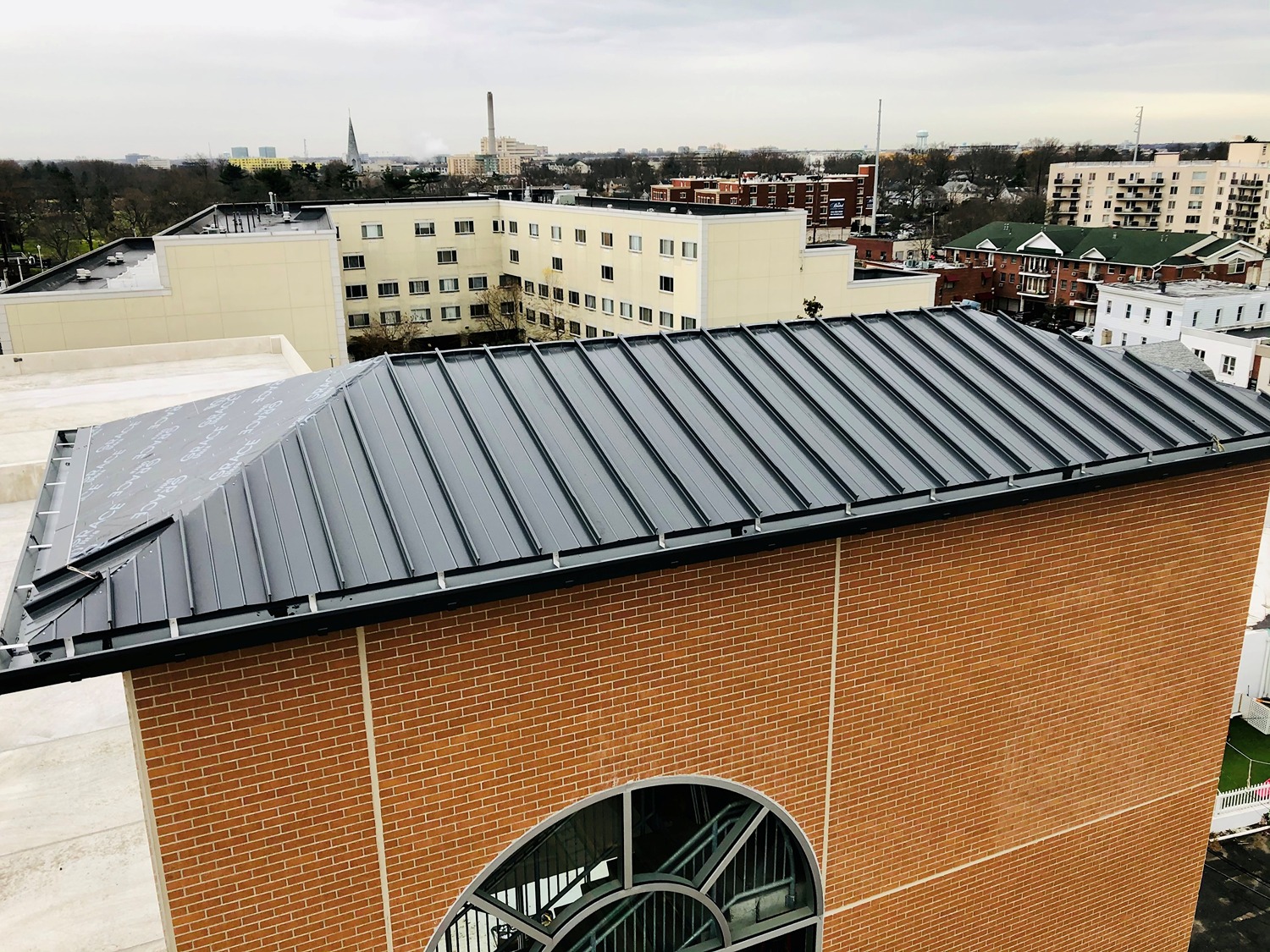 A newly installed dark gray metal standing seam roof atop a brick building with an arched window, overlooking a cityscape of varied buildings on a cloudy day.