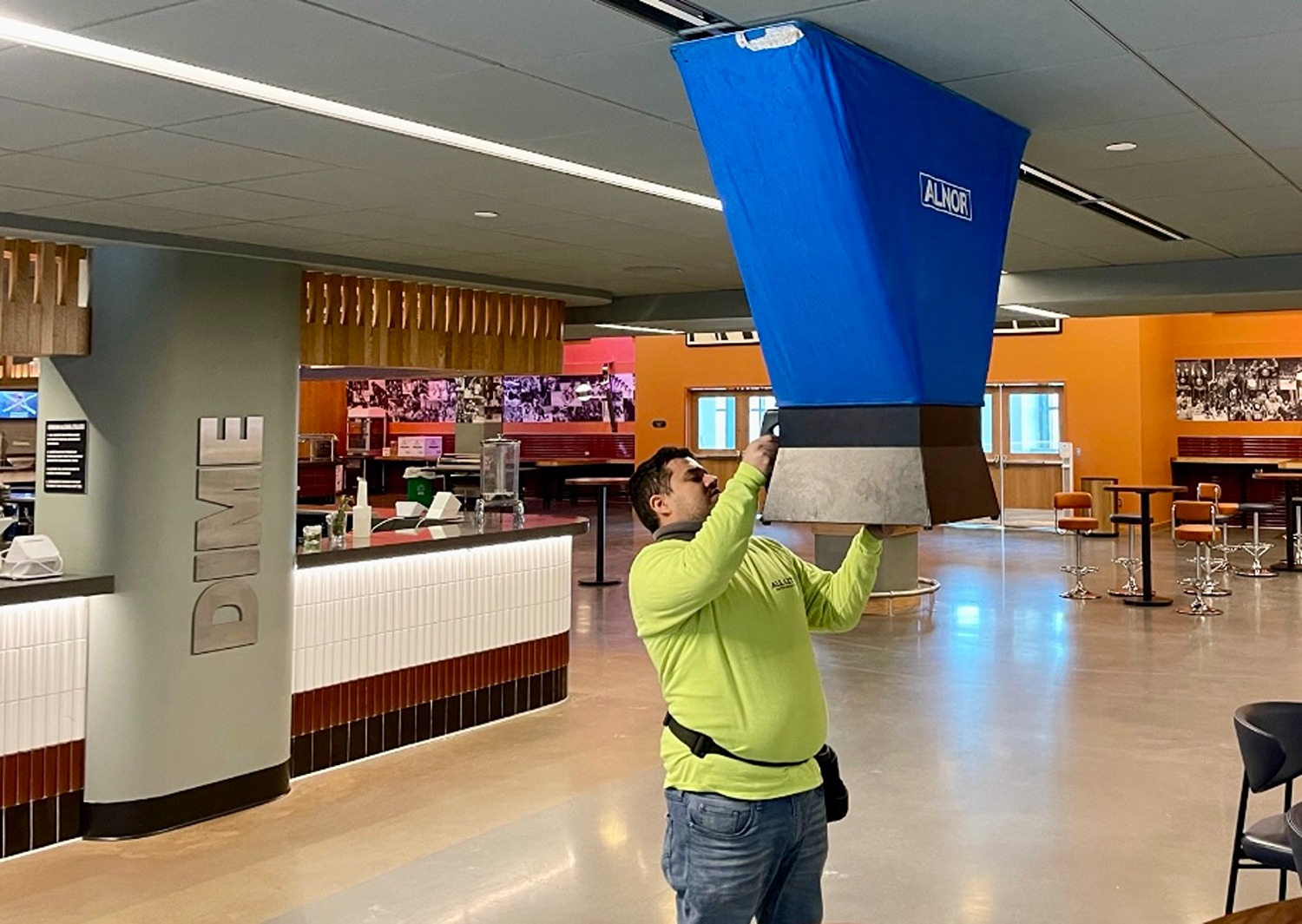 Man in a lime green shirt adjusting a large blue air duct labeled ALNOR in a modern indoor space with a reception desk and seating area.