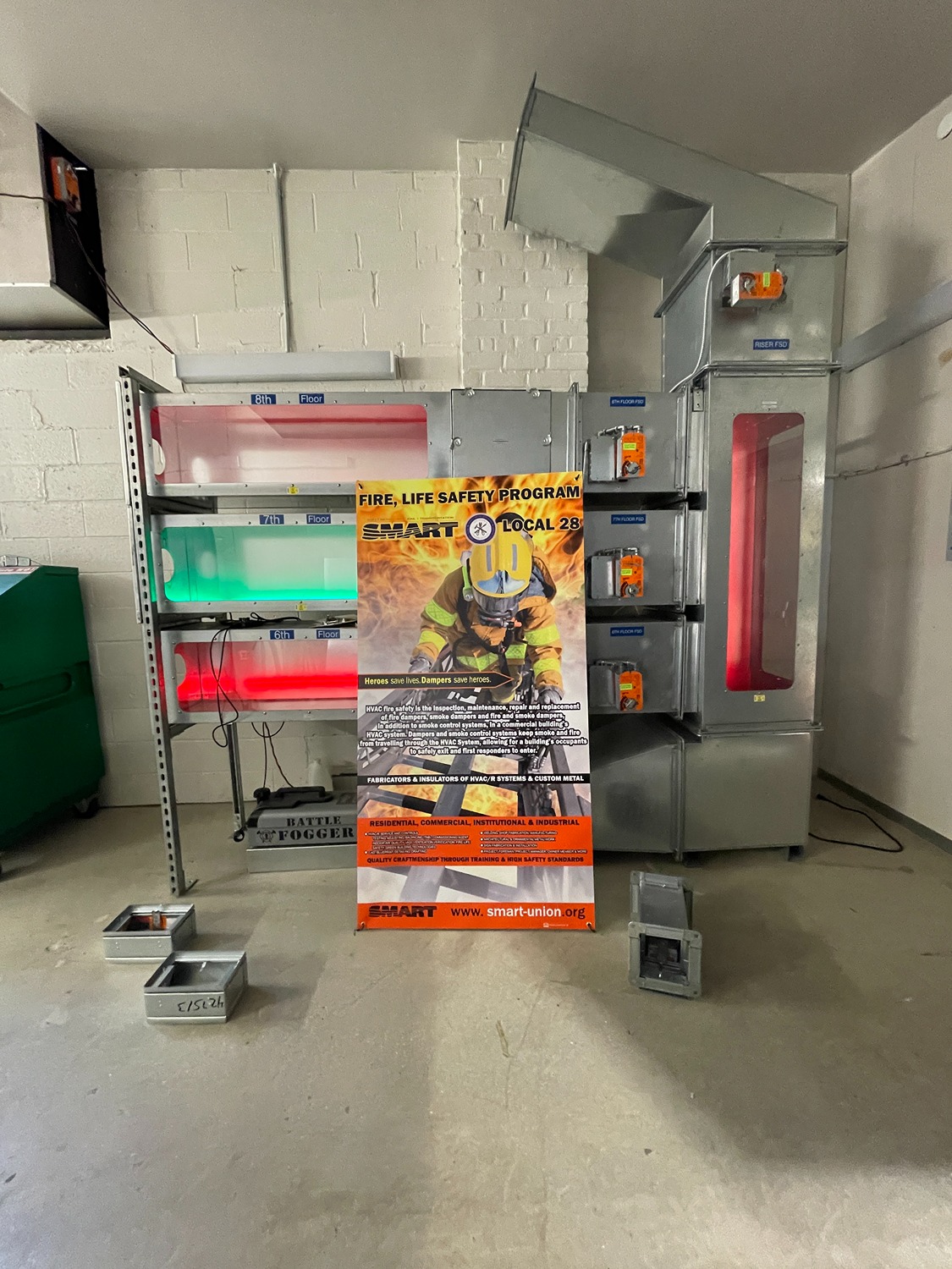 Industrial fire safety training setup with illuminated colored lights labeled by floor and a fire safety program poster featuring a firefighter.