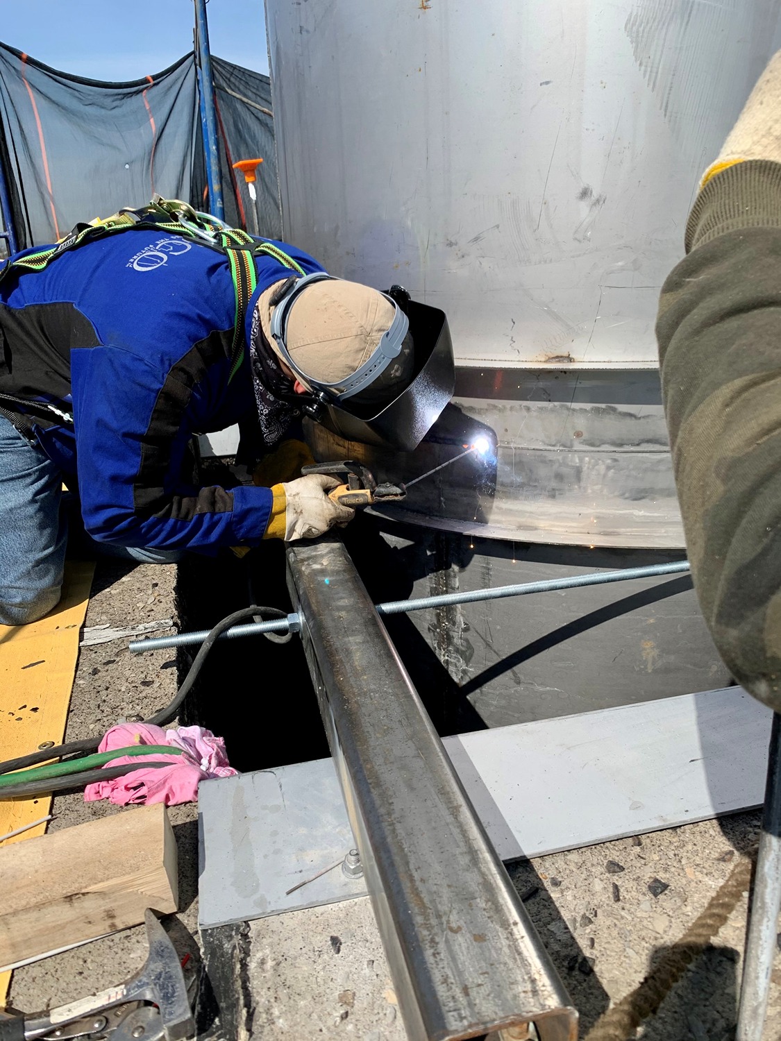 Worker in blue jacket and safety gear welding a metal pipe outdoors.