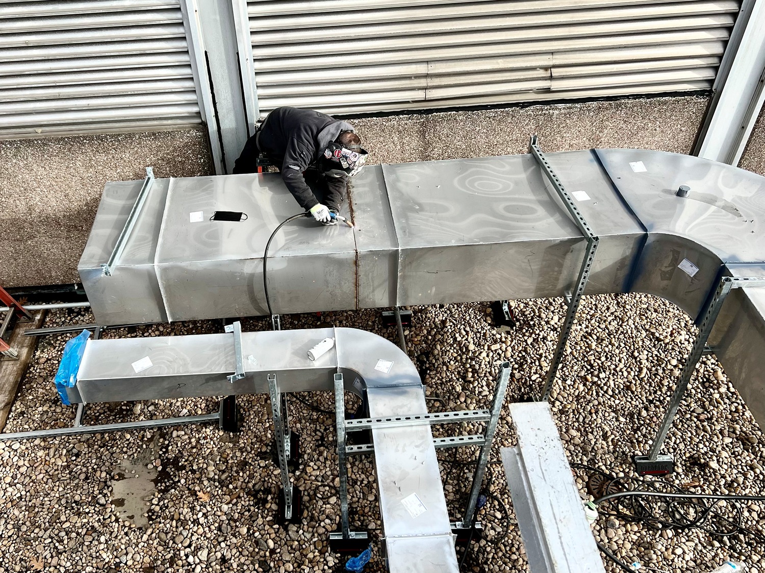 Worker wearing protective gear welding seams on large metal ductwork outdoors.