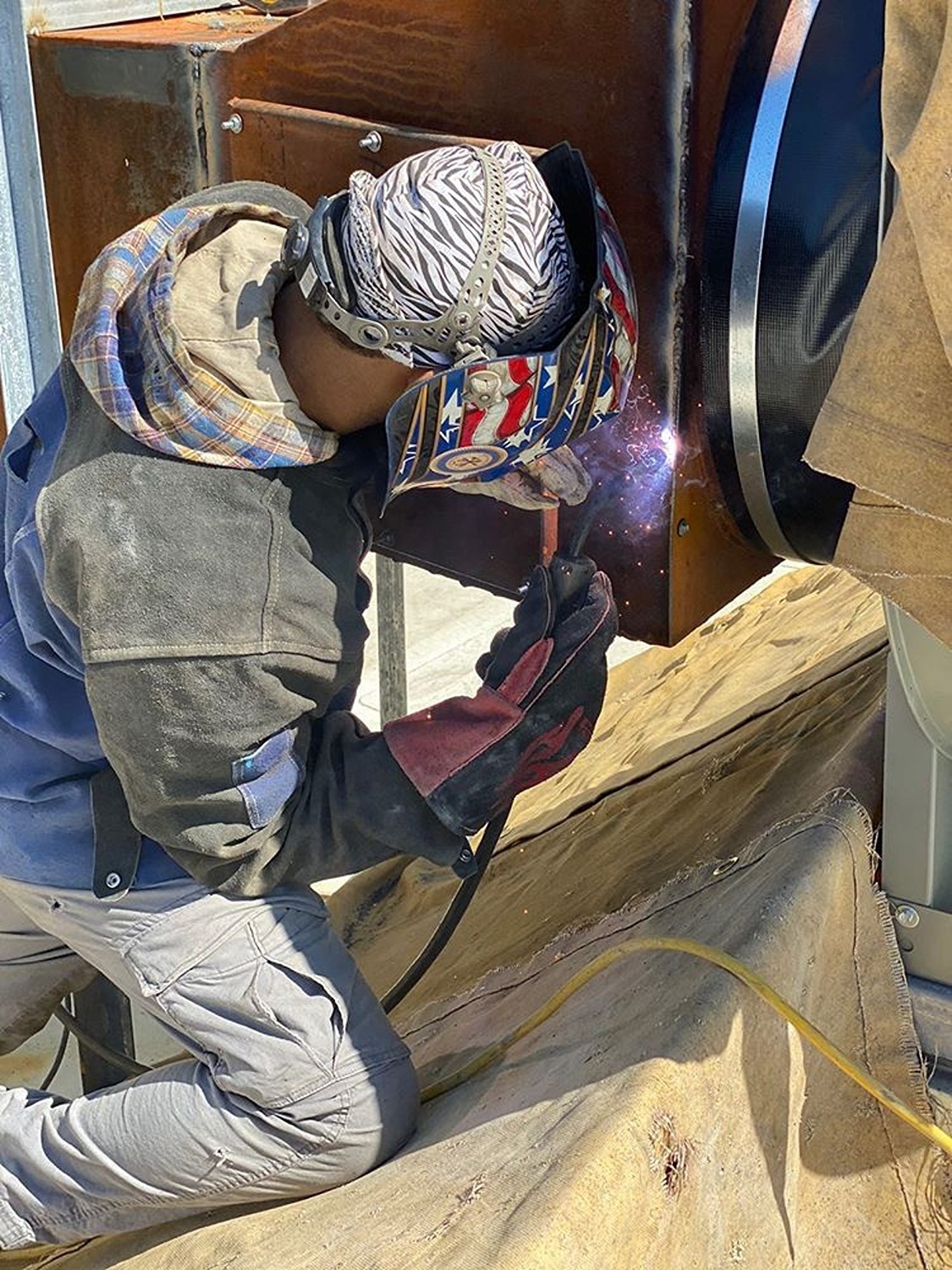 Welder wearing protective gear and a welding helmet with an American flag design working on a metal structure.