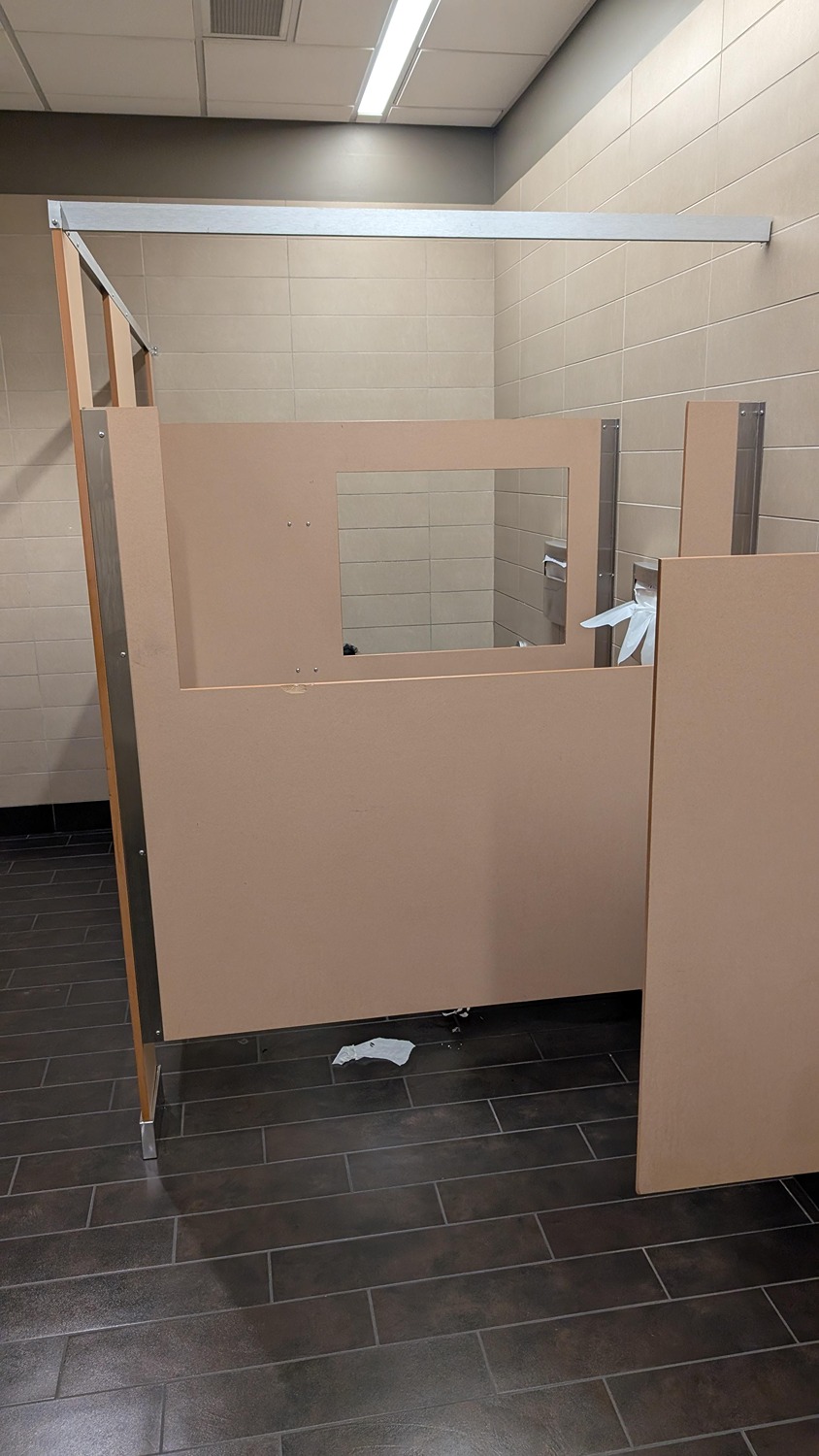 Beige-colored public restroom stall with an open door, a window cutout in the partition, and discarded paper on a dark tiled floor.