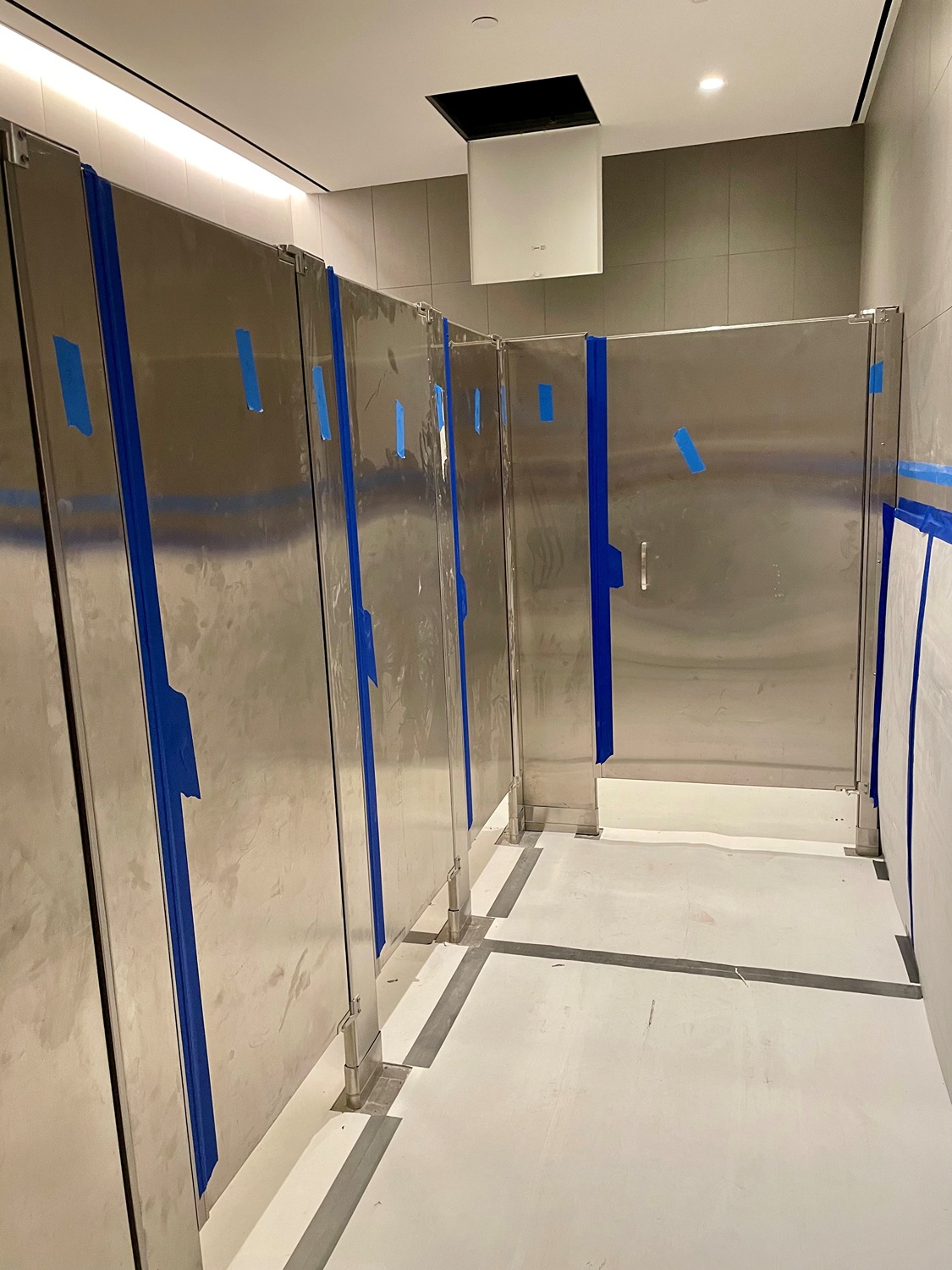 Row of stainless steel restroom stalls with blue painter's tape on doors and walls, under construction.