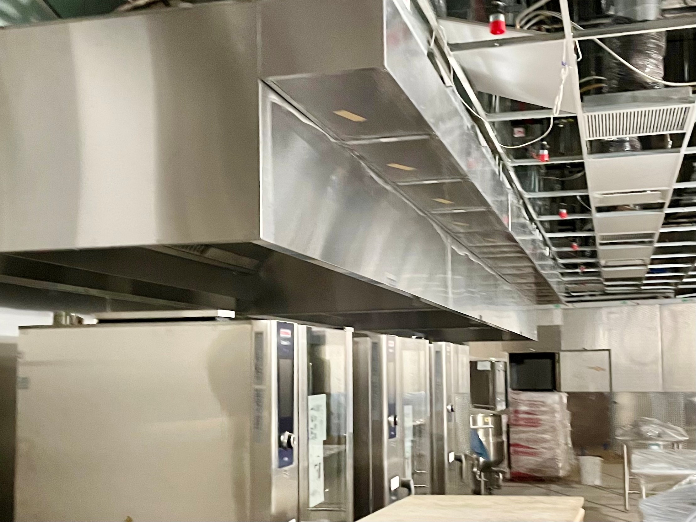 Commercial kitchen with stainless steel ovens beneath a large metal ventilation hood and an unfinished ceiling.