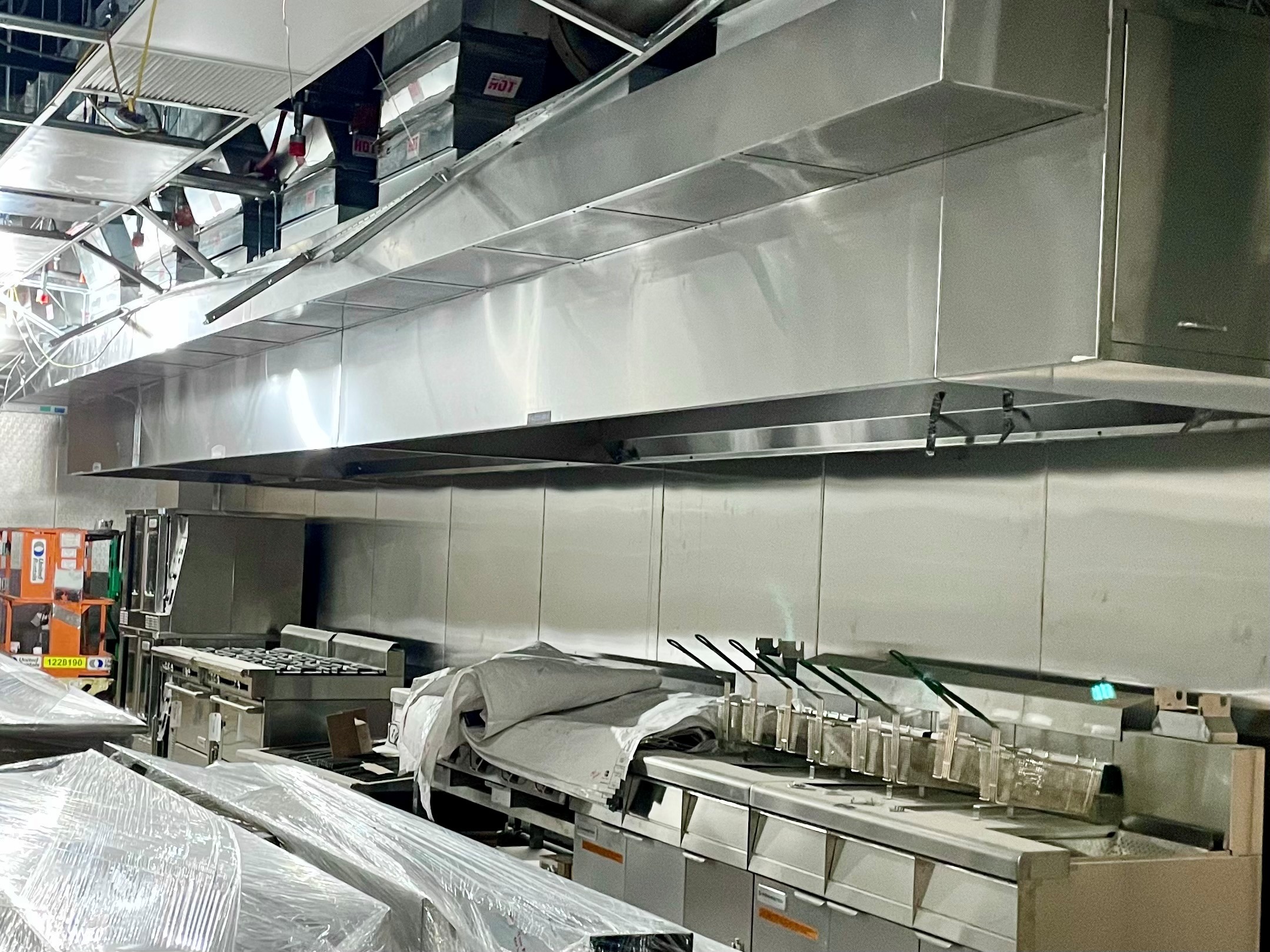 Industrial kitchen setup with large stainless steel ventilation hood, multiple stoves, deep fryers, and equipment covered in plastic wrap.