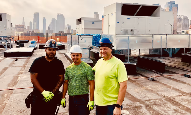Three construction workers wearing helmets and gloves standing on a rooftop with city buildings in the background.