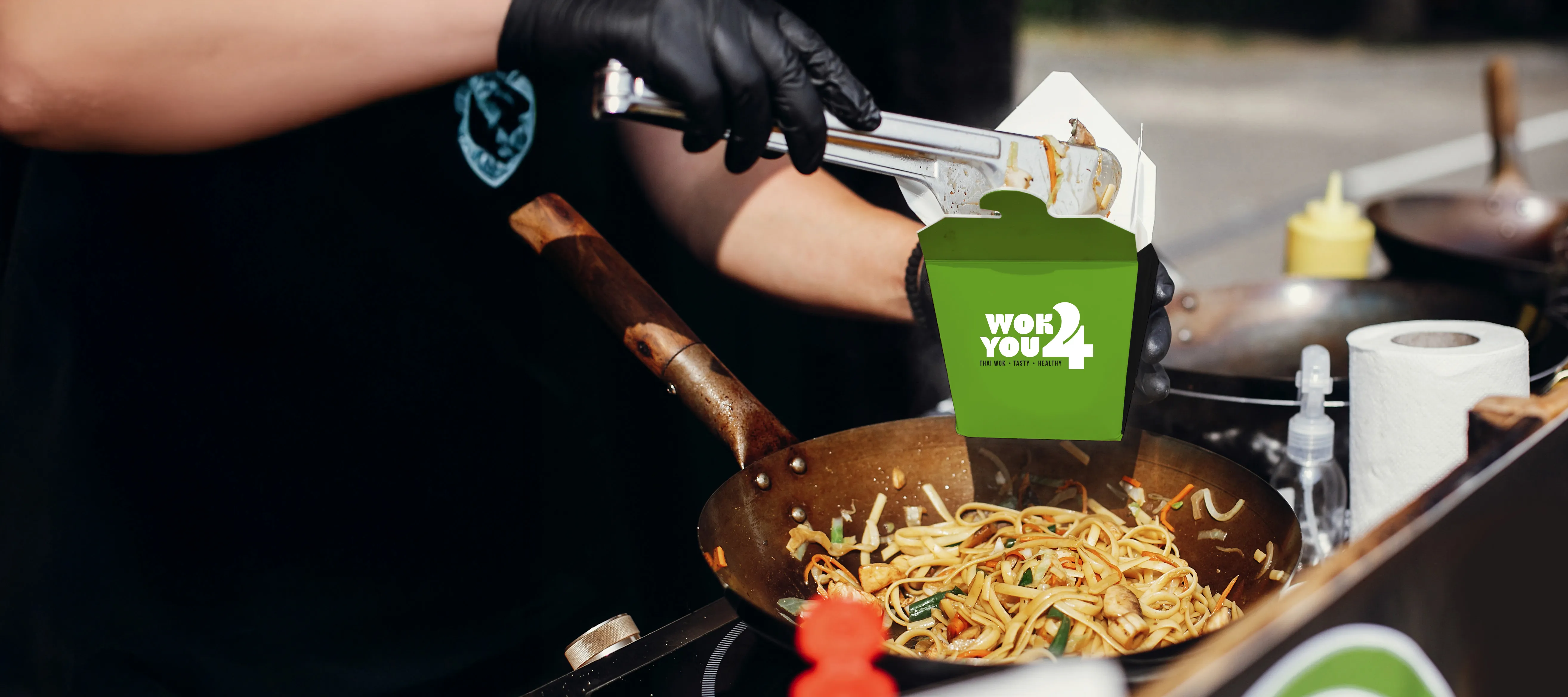 Chef wearing black gloves serving stir-fried noodles from a wok into a green Wok You 4 takeout box at an outdoor food stall