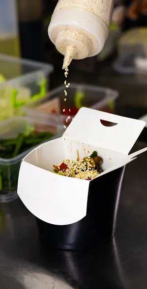 Sauce being drizzled over a takeout container filled with stir-fried vegetables and sesame seeds in a commercial kitchen setting.