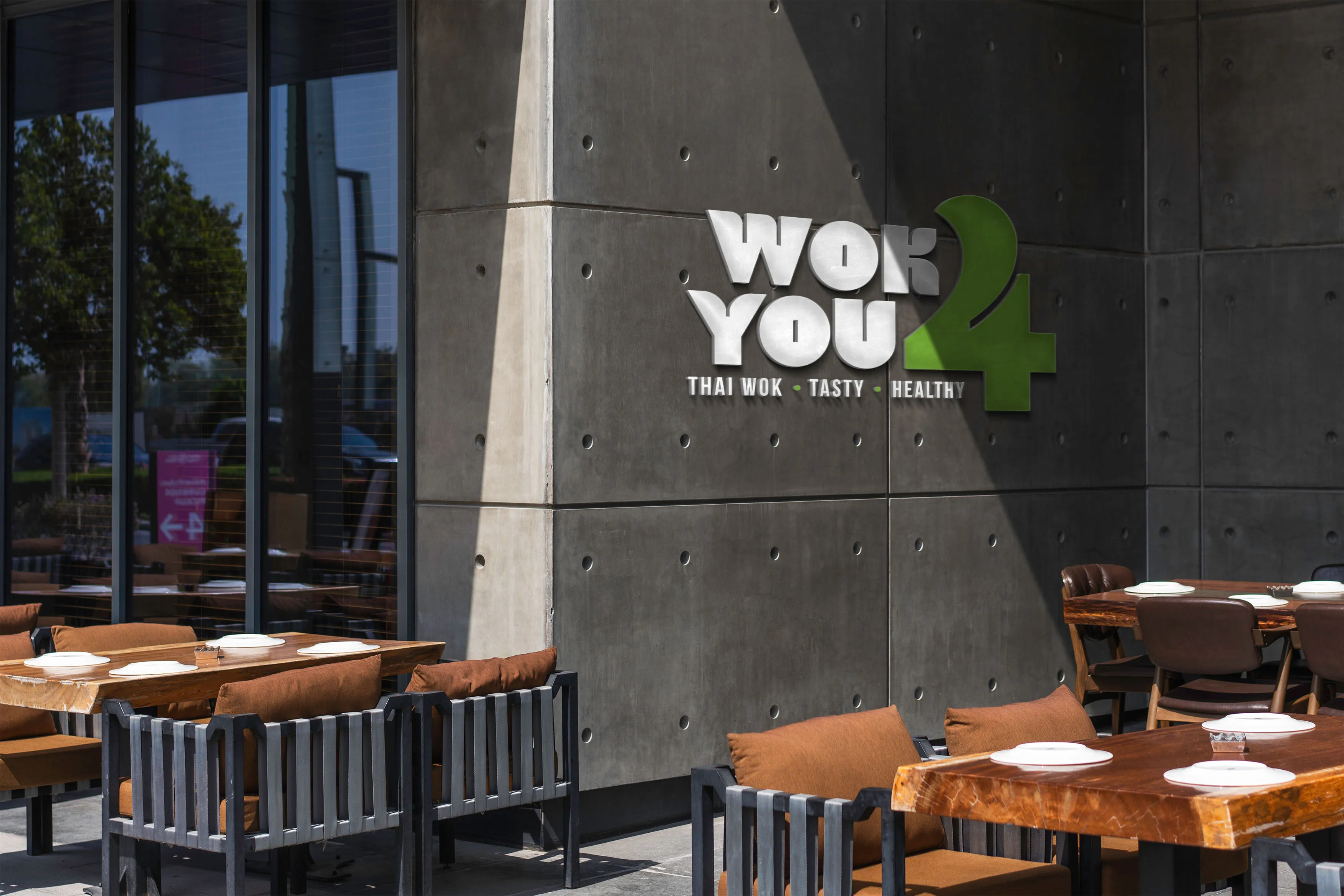 Outdoor seating area of Wok You 4 restaurant with modern wooden tables, cushioned chairs, and a concrete wall displaying the restaurant’s logo and slogan: Thai Wok • Tasty • Healthy