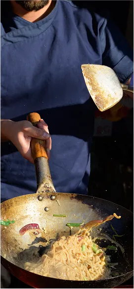 Chef en uniforme bleu faisant sauter des nouilles avec des légumes dans un wok, capturé en pleine action avec des ingrédients qui volent.
