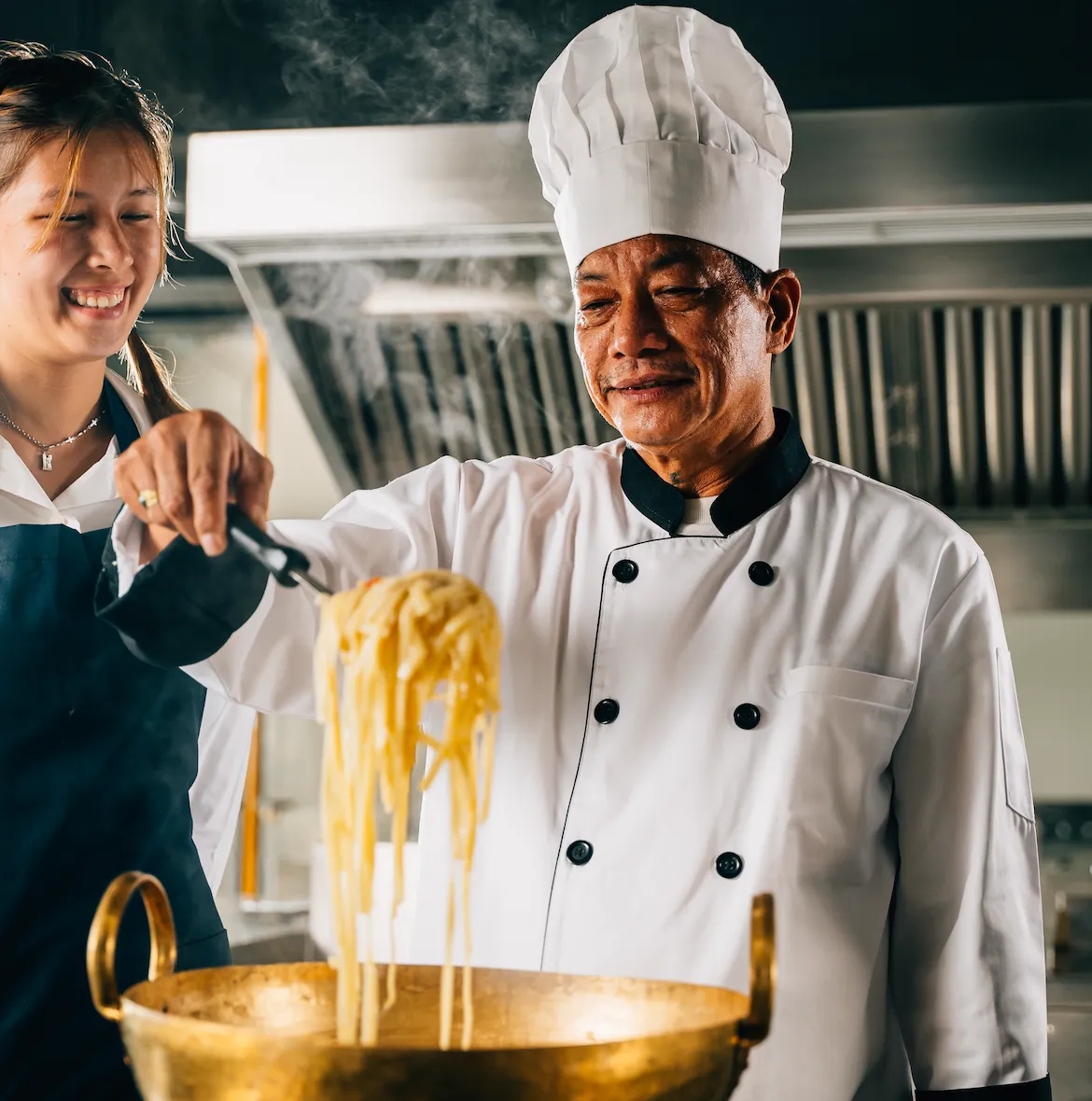 Smiling chef in a white uniform and hat lifting freshly cooked noodles from a pot with tongs, accompanied by a young assistant in a professional kitchen