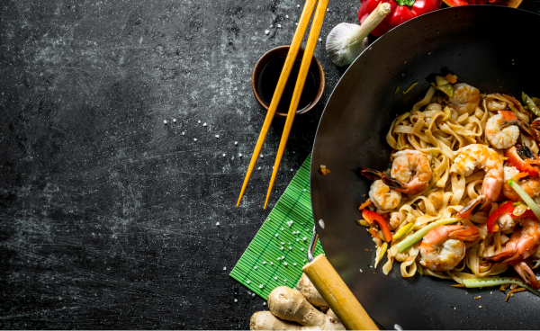 Wok rempli de nouilles, de crevettes et de légumes sautés sur une surface sombre, avec des baguettes, de la sauce soja, du gingembre et des ingrédients frais à proximité.