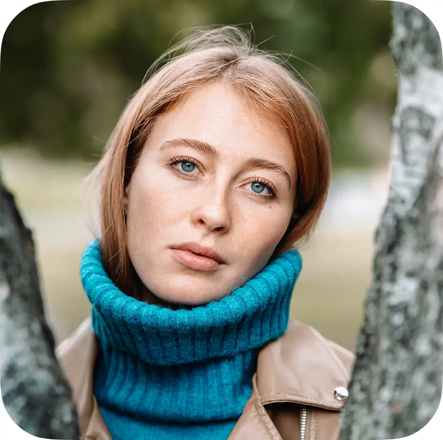 A person wearing a teal turtleneck and a beige jacket stands between two trees, with a blurred face. The background is softly out of focus.