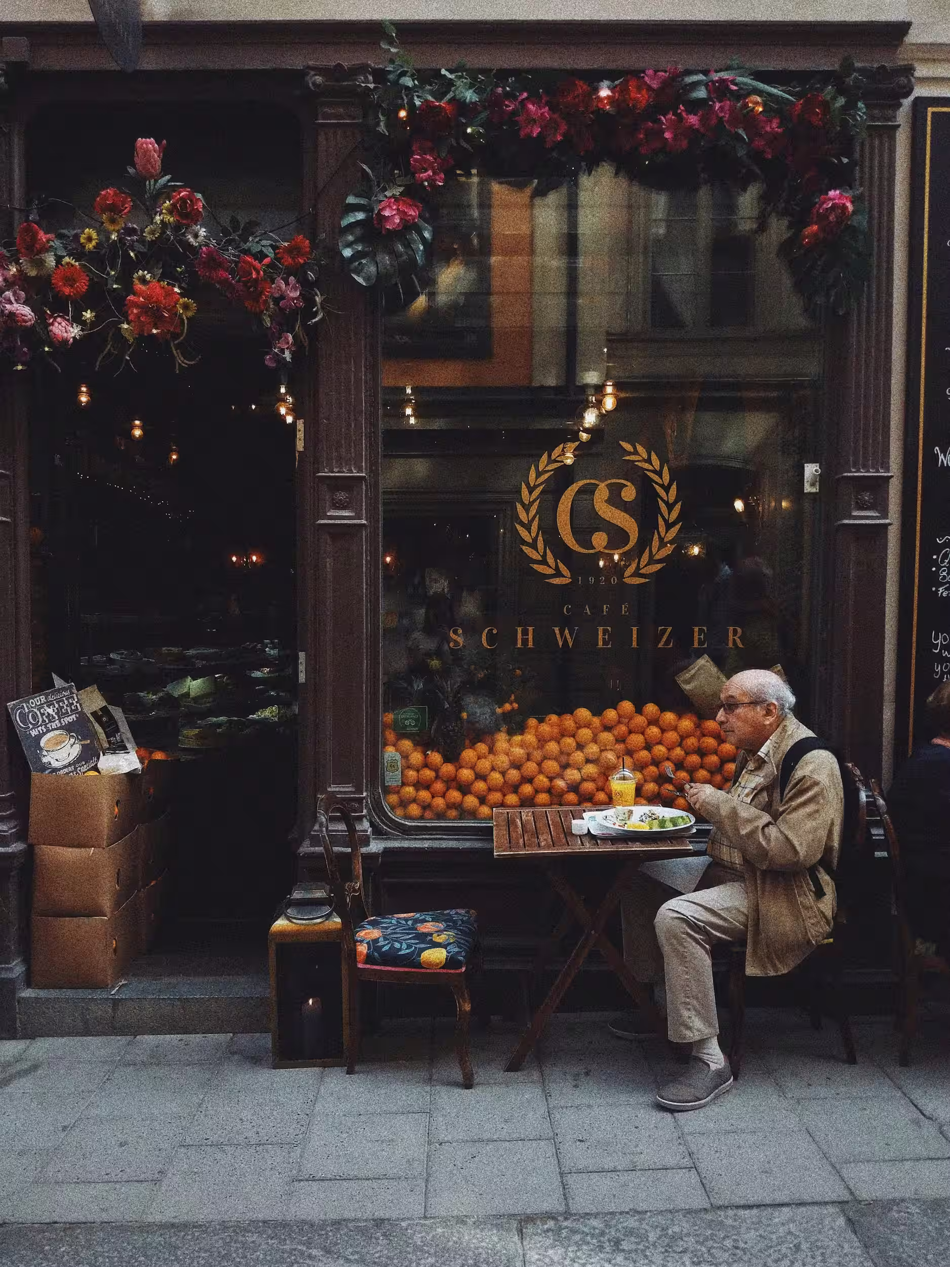 Man in Brown Jacket Sitting on Chair in Front of Fruit Stand.