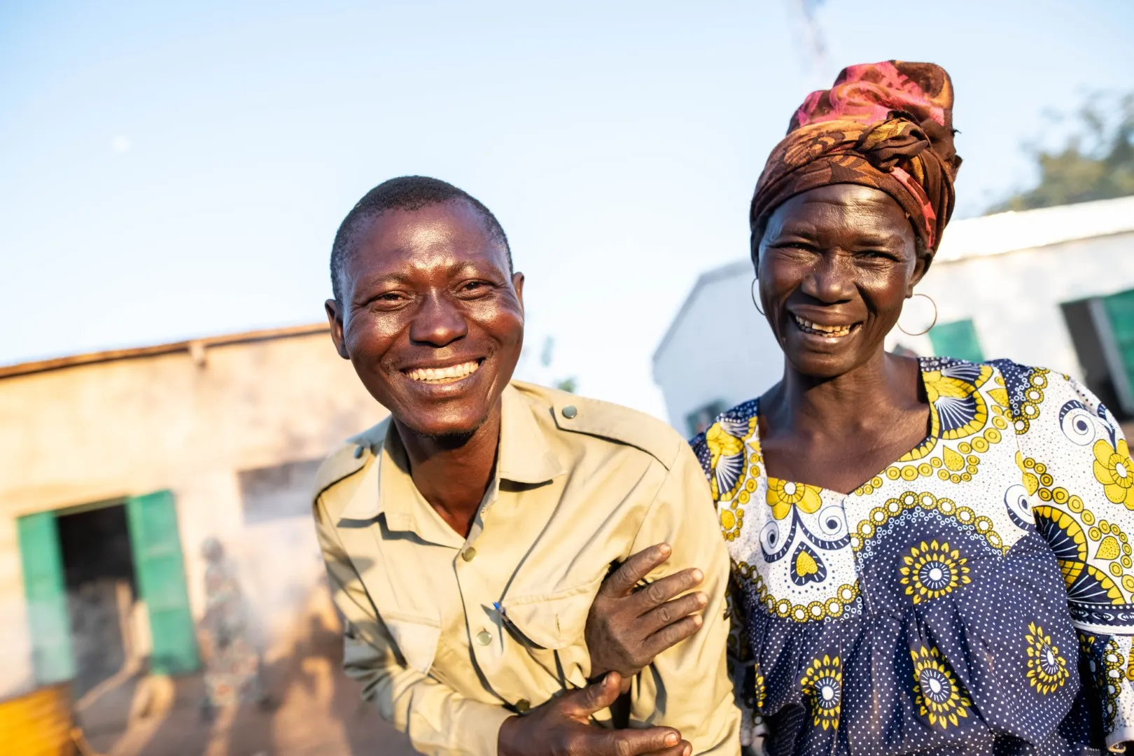 Un homme et une femme souriants, debout dehors devant des bâtiments au soleil.