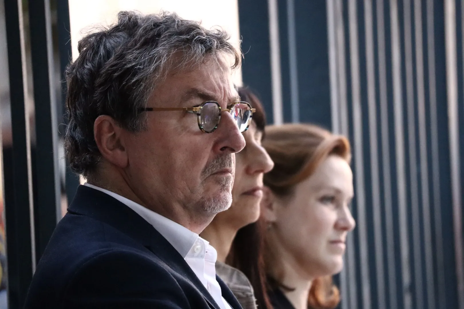 Profil de trois adultes regardant attentivement vers la droite sur un fond de barres verticales.