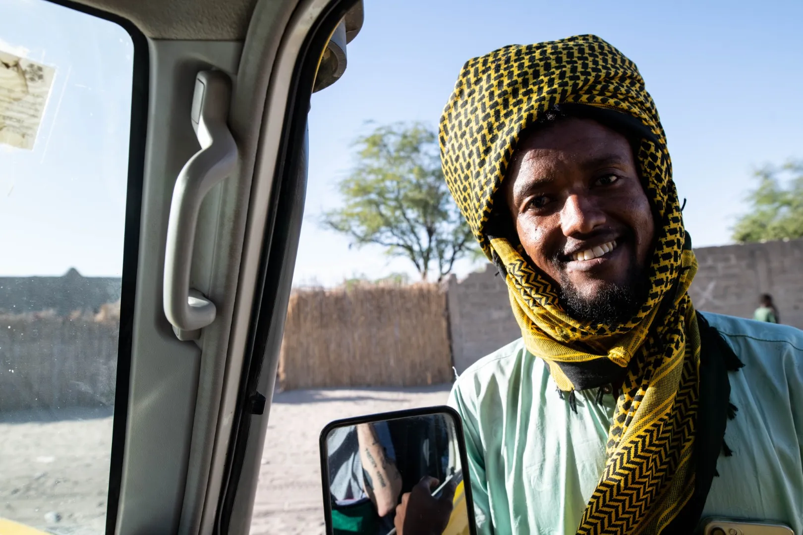 Un homme souriant porte un foulard jaune à motifs noirs, debout à l'extérieur près d'un véhicule.