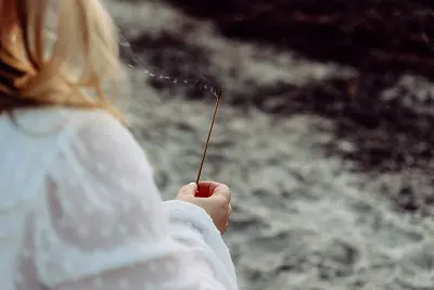 Eine Person in Weiß hält ein Räucherstäbchen in der Nähe von Wasser, von hinten gesehen.