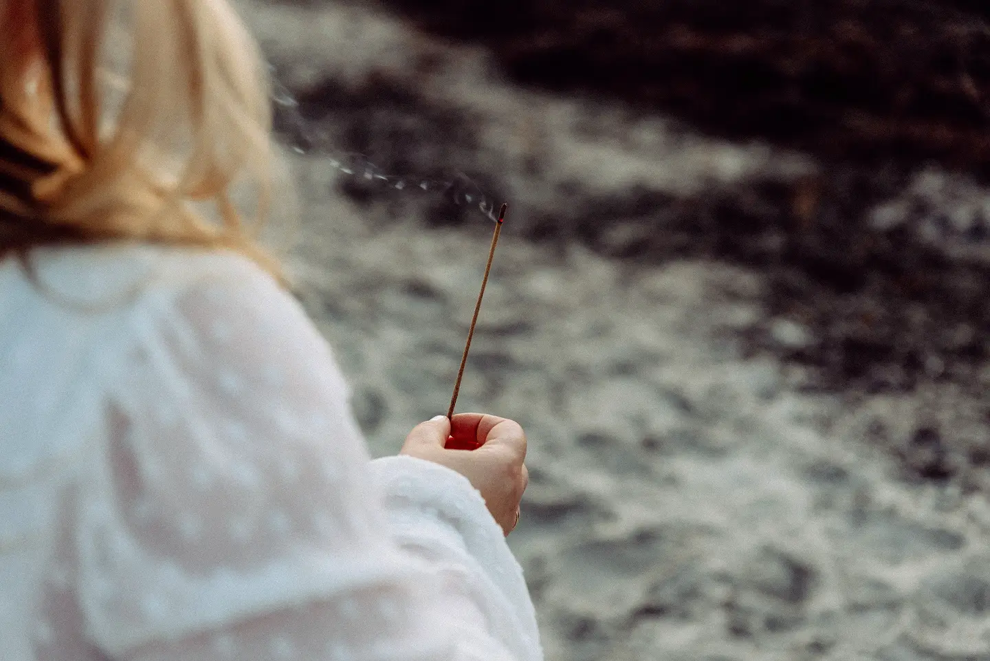 Eine Person in Weiß hält ein Räucherstäbchen in der Nähe von Wasser, von hinten gesehen.