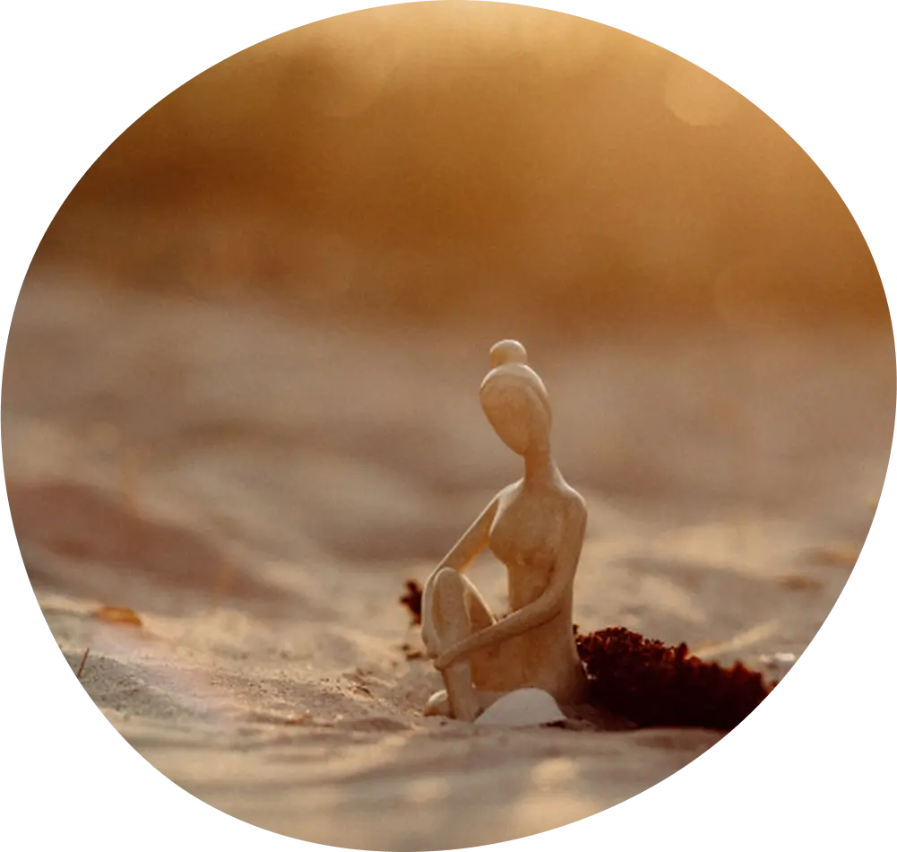 Eine kleine Skulptur einer sitzenden Figur auf sandigem Boden, die in warmes, goldenes Sonnenlicht getaucht ist.