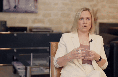 Woman in a light-colored suit speaking and gesturing while seated in a modern indoor setting.
