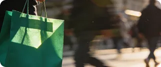 Person carrying a green shopping bag walking outdoors with blurred pedestrians in background.